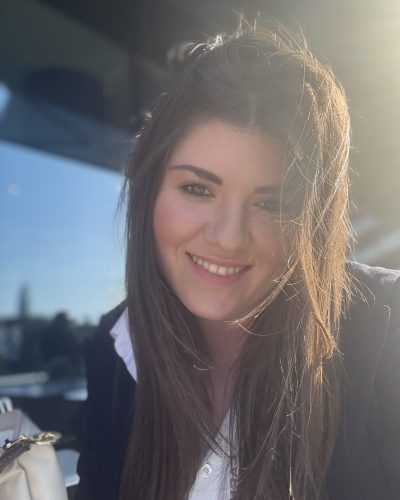 Close-up portrait of a smiling woman with long brown hair, illuminated by warm sunlight on one side.